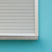 Corner of window covered by metal security blinds, framed by a light blue wall.